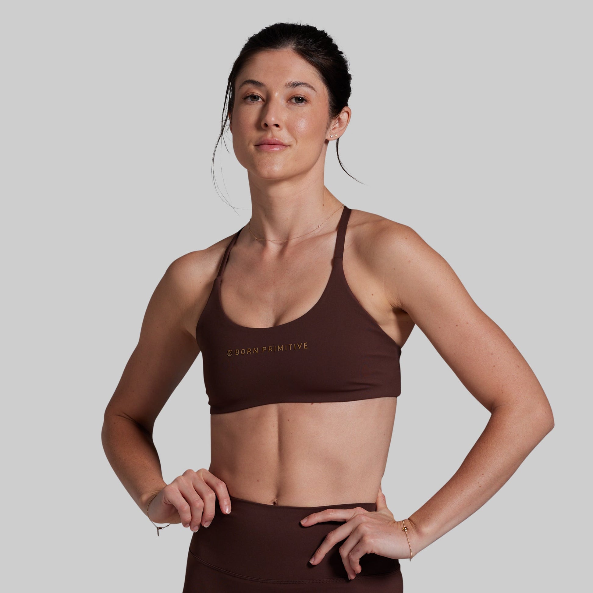 A woman modeling a dark brown scoop-neck sports bra with gold 'BORN PRIMITIVE' branding on the chest, paired with matching brown athletic bottoms, against a solid light gray background.