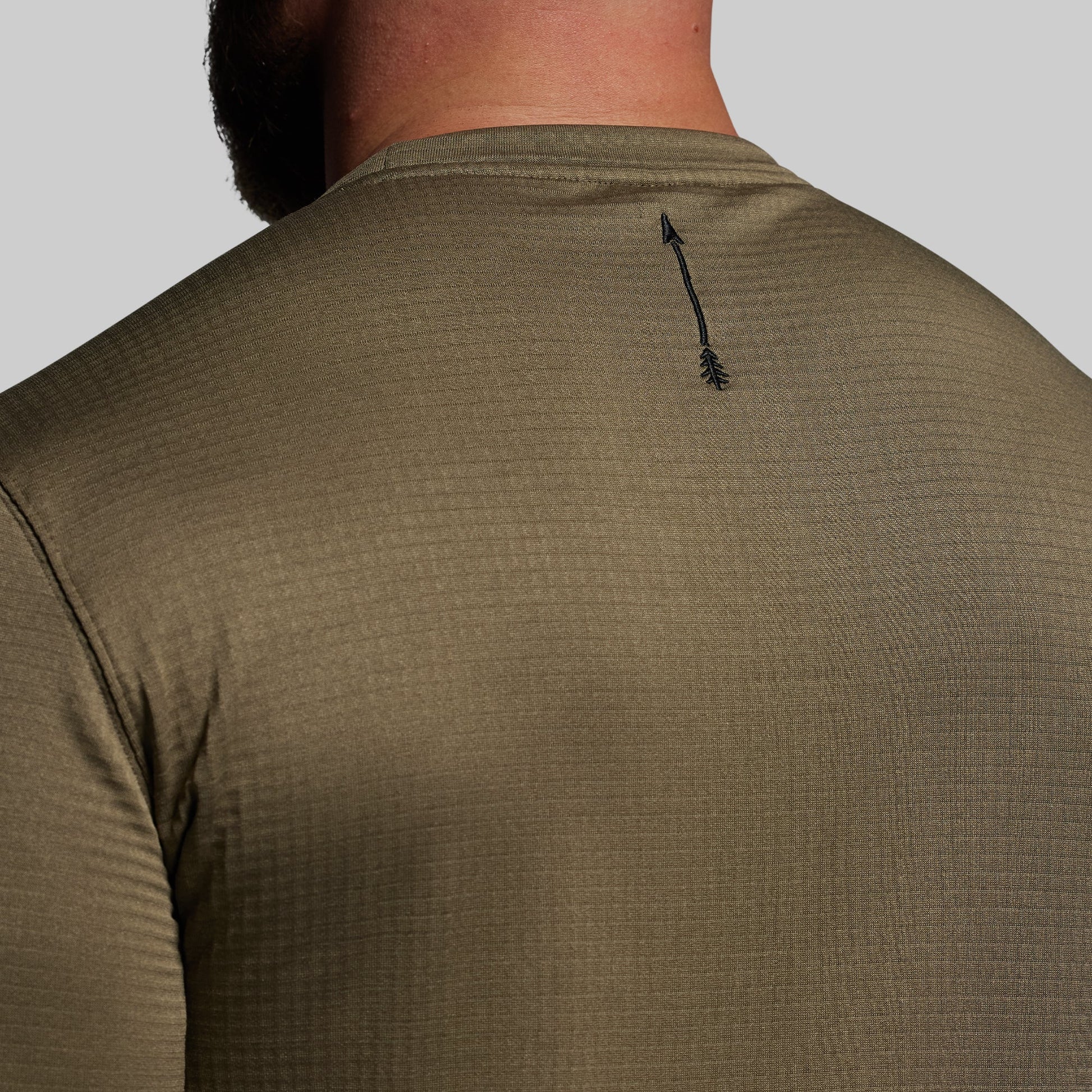 Close-up of a person wearing a green textured shirt with a small black logo on the back against a gray background
