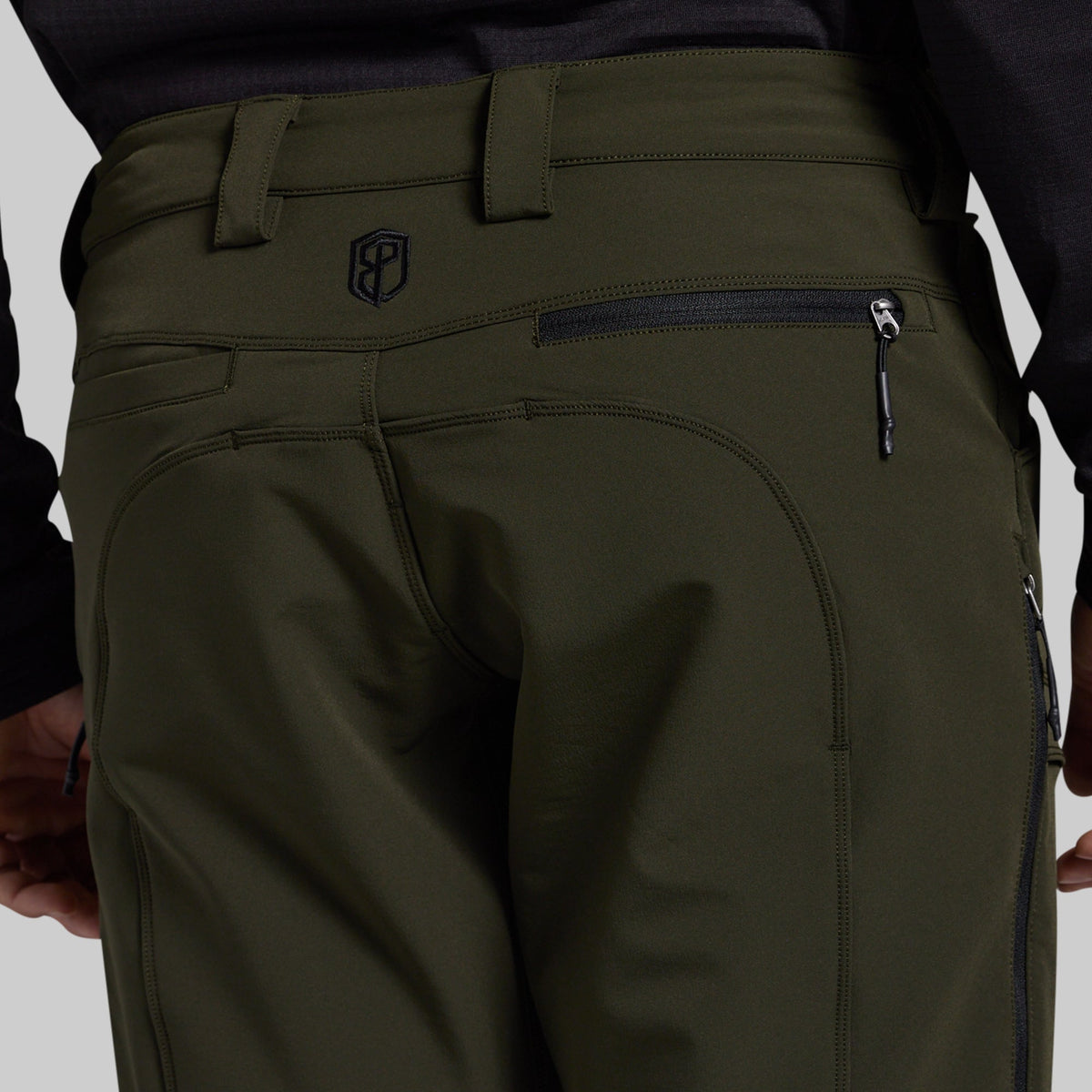 Close-up rear view of olive green outdoor pants worn by a model, showing the back waistband, belt loops, zippered pocket, and a small shield logo near the upper left side.