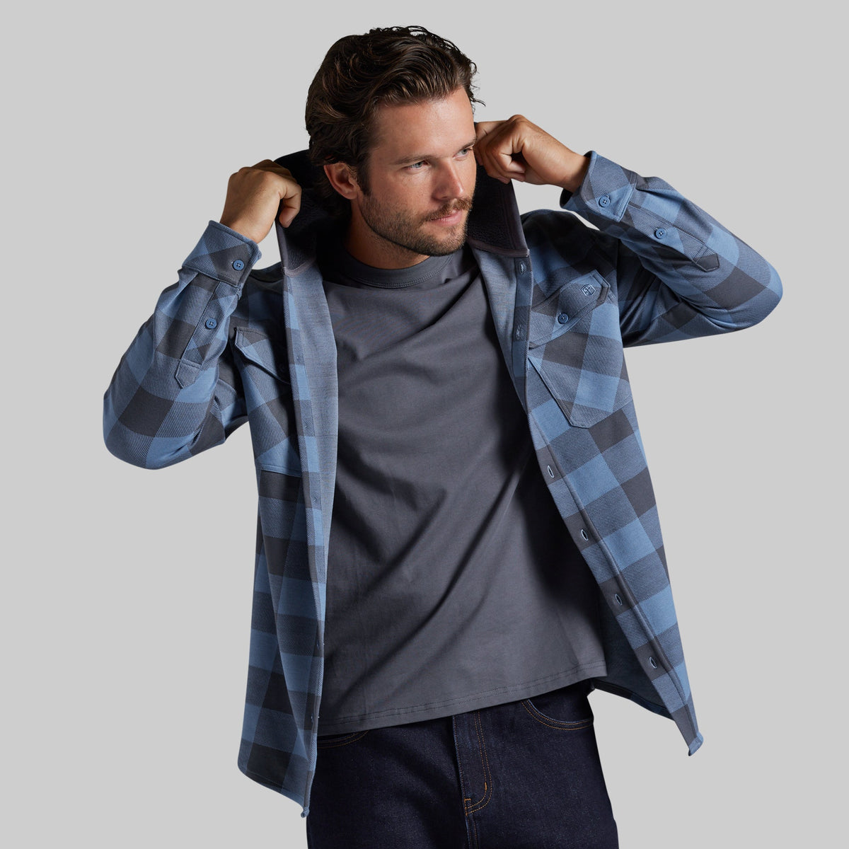 A man wearing a blue and gray plaid hooded flannel shirt over a dark gray t-shirt and dark jeans.