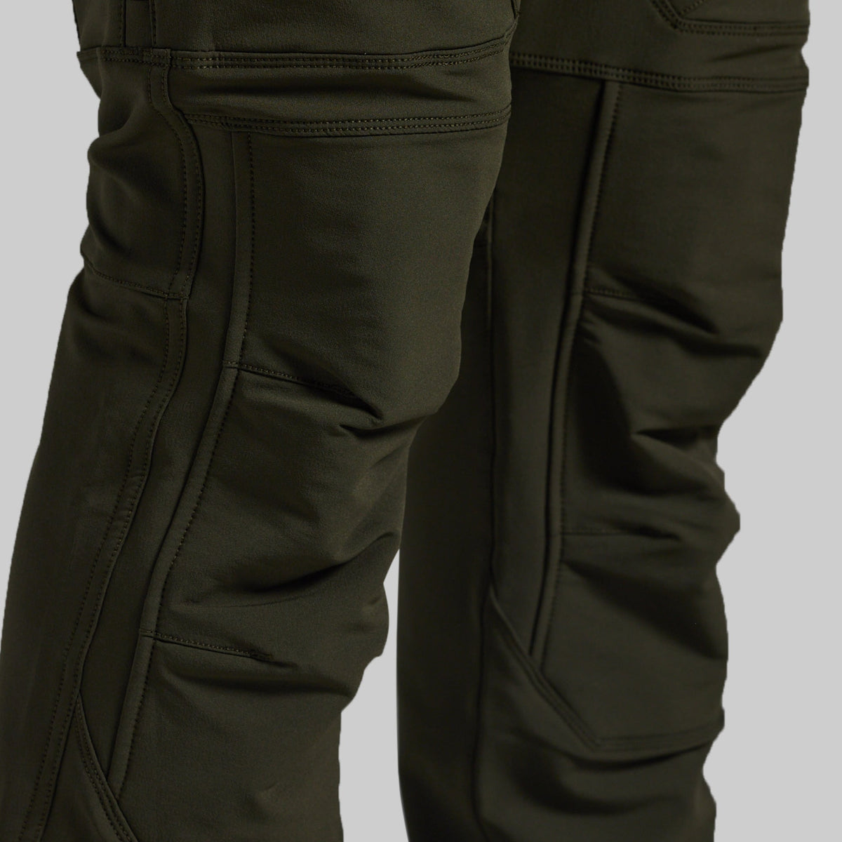 Close-up of dark olive green technical pants showing the leg construction, seam details, and pocket stitching against a neutral gray background.