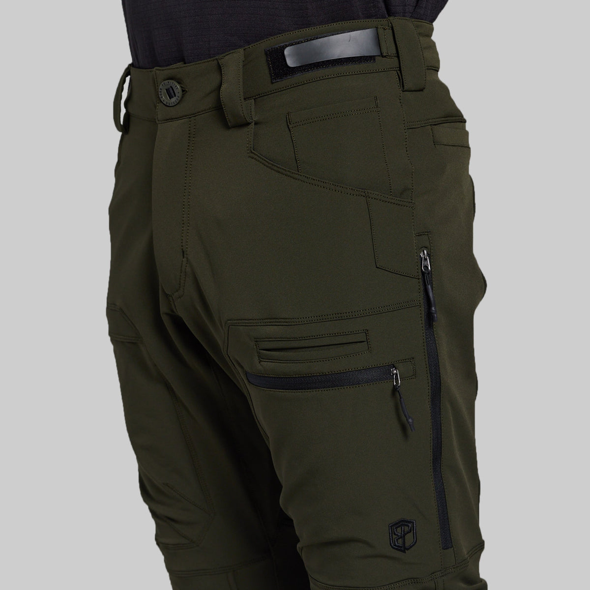 Close-up of olive green technical pants with multiple zippered cargo pockets, belt loops, and a dark belt, shown on a model against a light gray background.