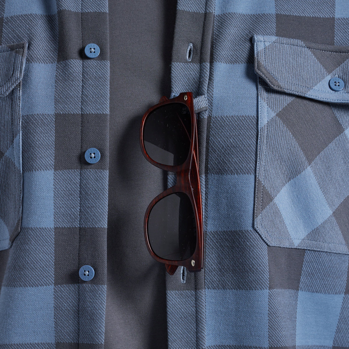 A pair of sunglasses tucked into the sunglass loop of a gray and blue plaid flannel shirt.
