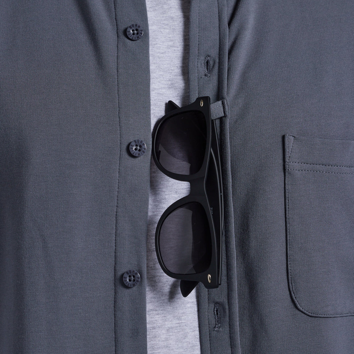 Black sunglasses shown hanging from a pocket strap on a dark gray button-up shirt over a light gray t-shirt.