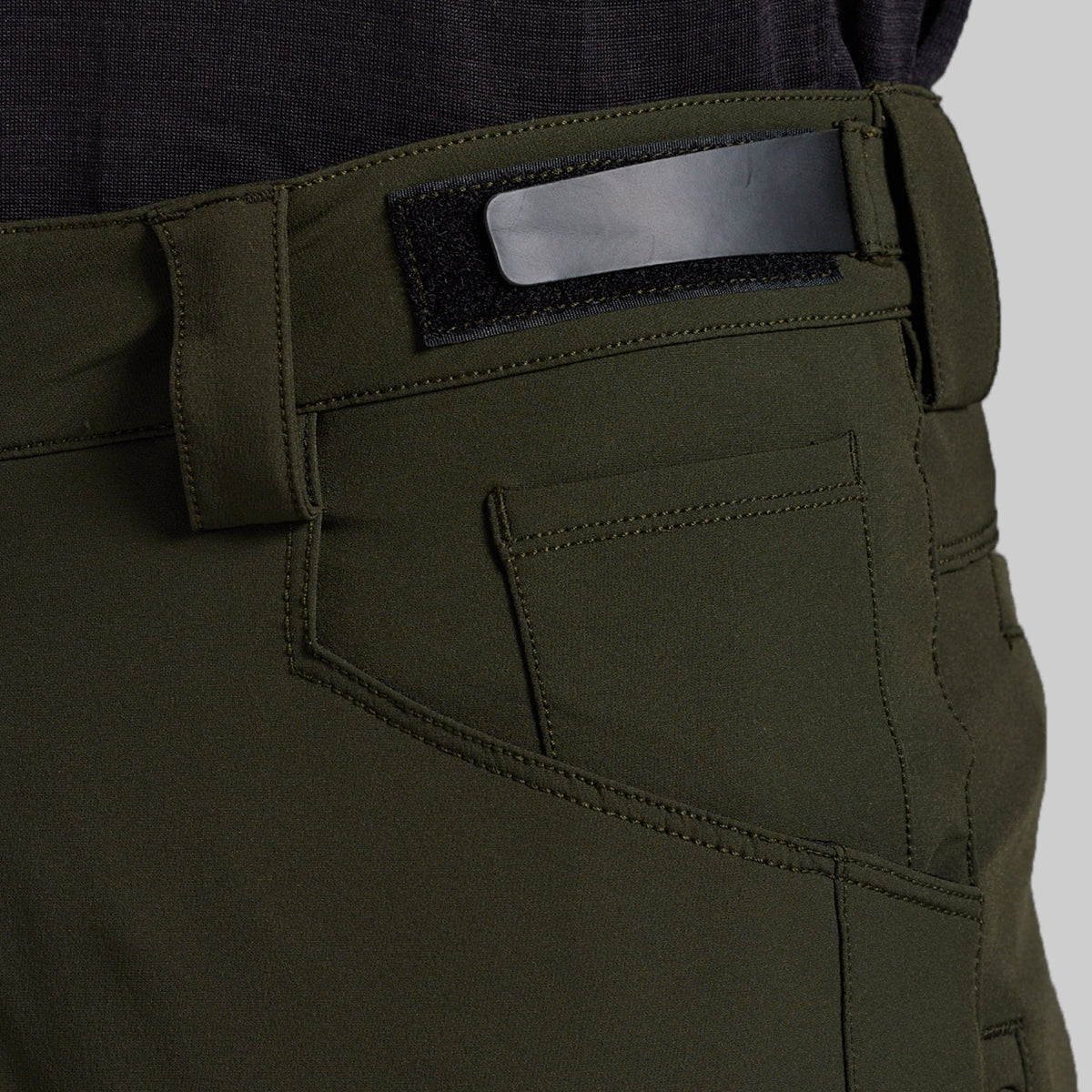 Close-up of the back waistband and pocket area of olive green outdoor pants, showing a black hook-and-loop patch with a silver metal bar, stitching, and belt loops.