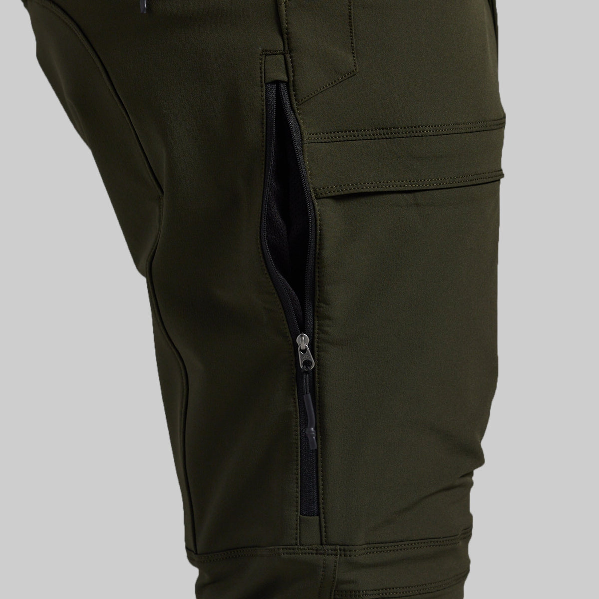 Close-up of olive green pants showing a side cargo pocket with a vertical zipper and a flap pocket above it, photographed against a light gray background.