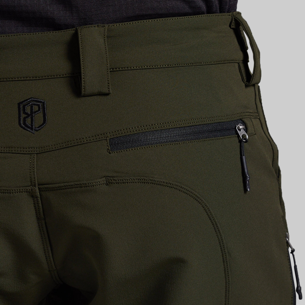 Close-up of olive green outdoor pants showing a zippered pocket, belt loop, and a black embroidered logo on the back.