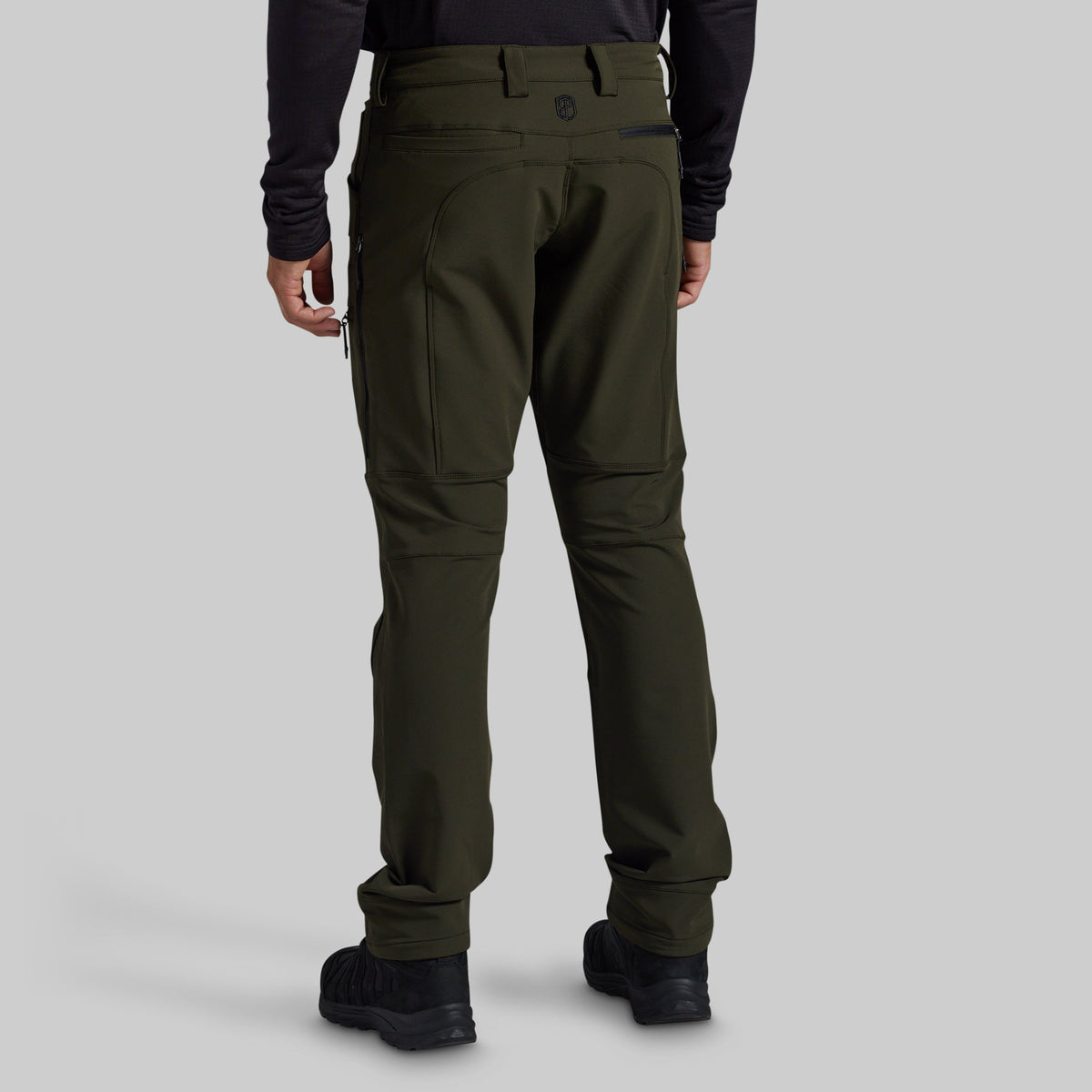 Rear view of a person wearing dark olive green technical pants with a black long-sleeve top and black shoes, shown against a light gray studio background.