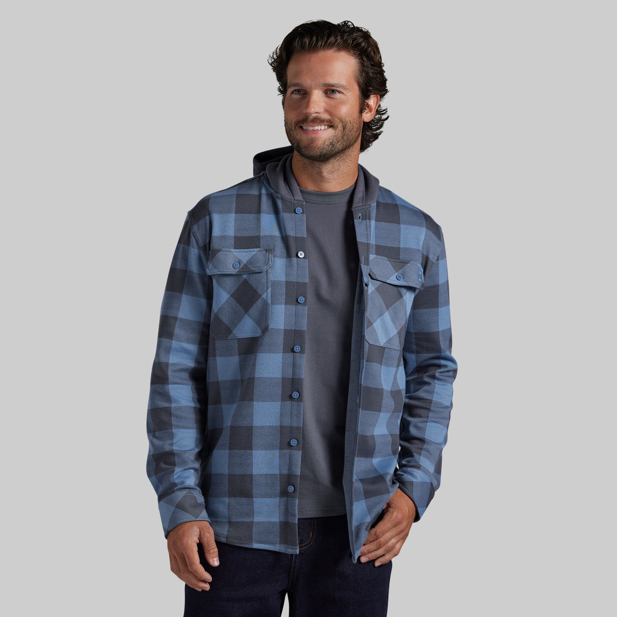 A man wearing a blue and gray plaid hooded button-up shirt over a dark gray t-shirt and dark jeans, standing against a solid gray background.