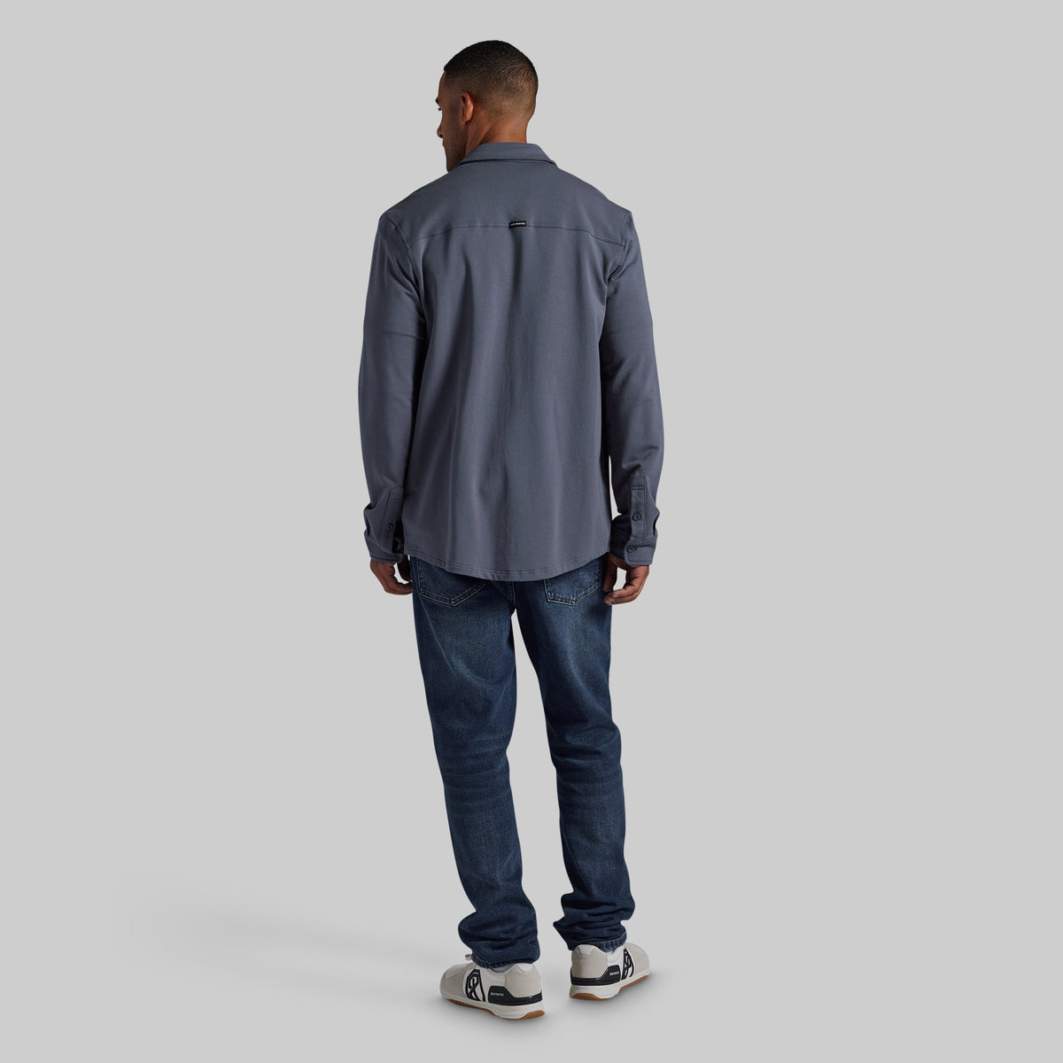 A man viewed from behind wearing a slate gray long-sleeve button-up shirt, dark blue jeans, and white sneakers, standing against a solid light gray background.