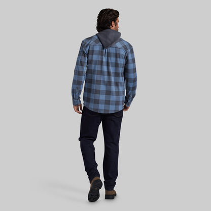 A man viewed from behind wearing a blue and gray plaid hooded flannel, dark navy pants, and brown shoes, standing against a solid light gray studio background.