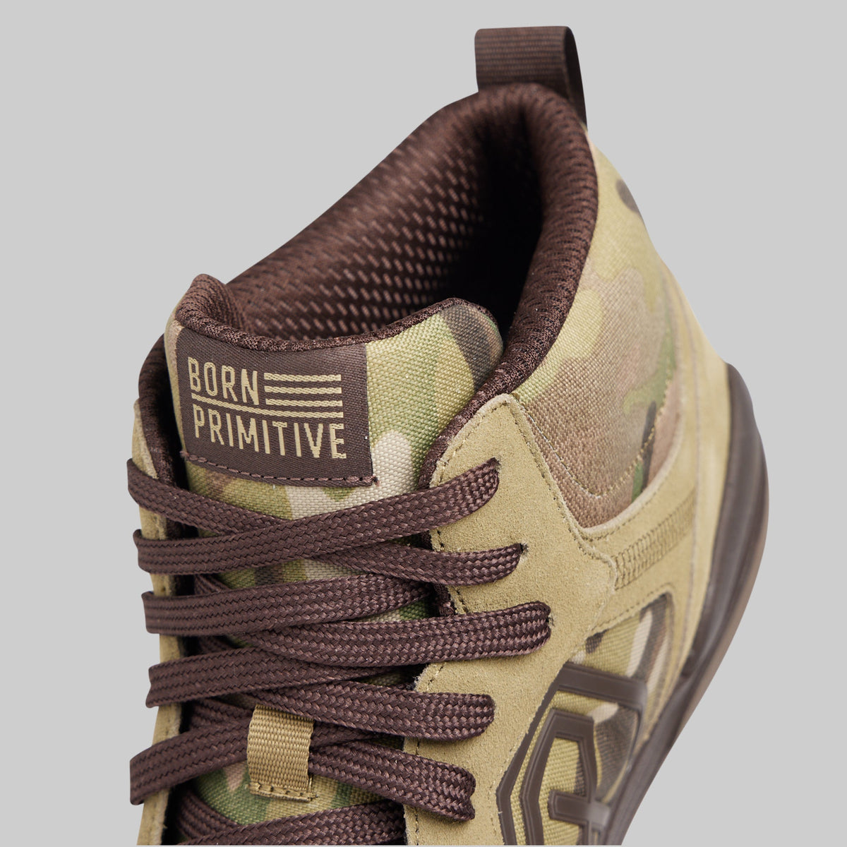 A close-up shot of a mid-top sneaker by Born Primitive, featuring a tan and olive green camouflage pattern, dark brown laces, and a brown tongue label with the brand name.