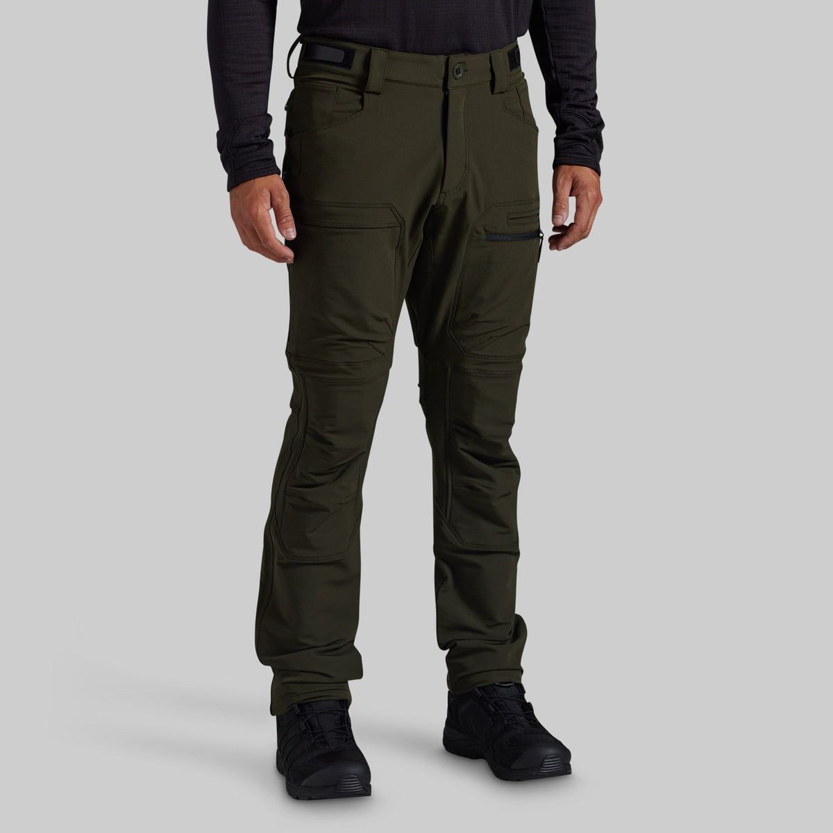 A person wearing dark olive green outdoor cargo pants with black boots and a black long-sleeve shirt, shown from the waist down against a light gray studio background.