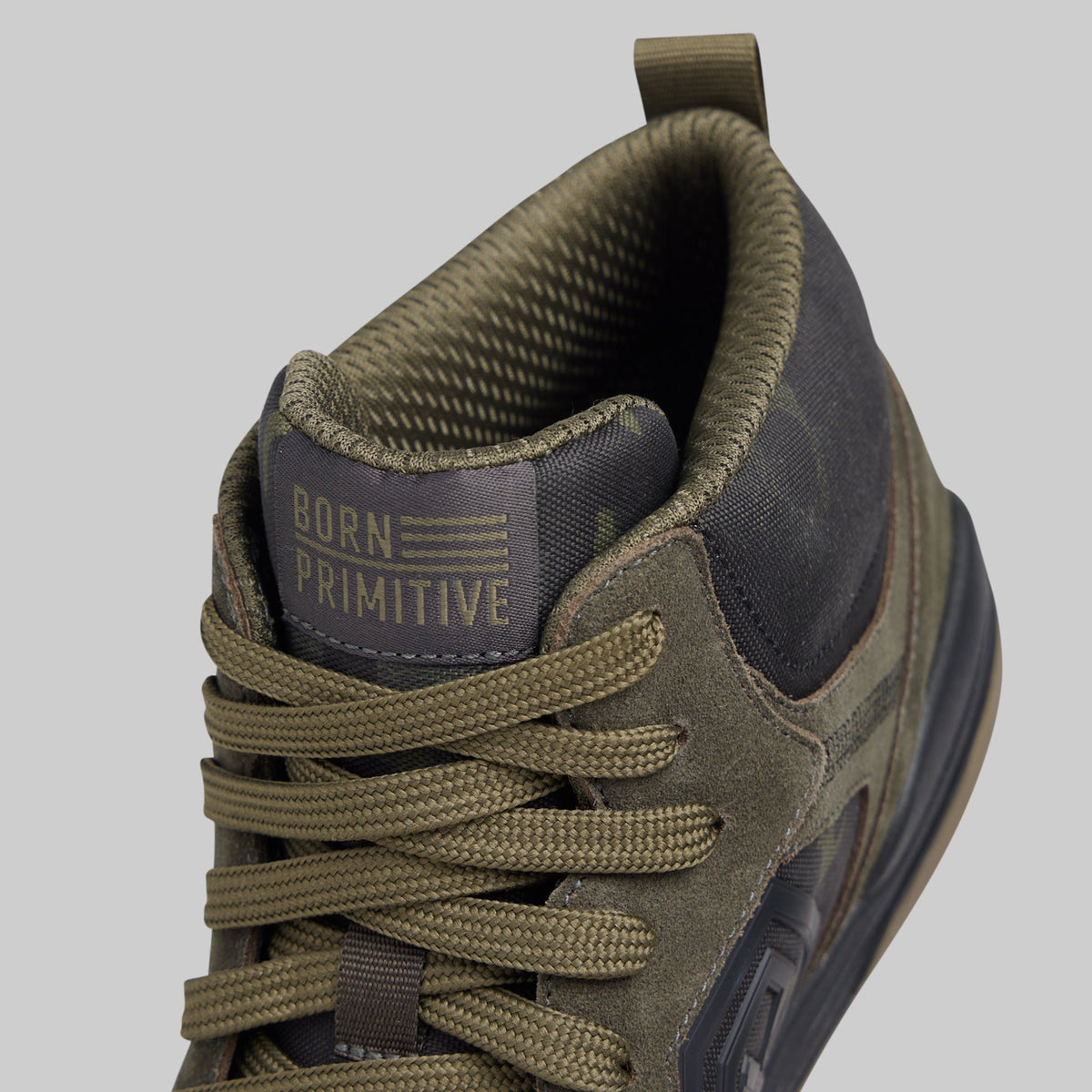 Close-up of a mid-top sneaker in olive green and dark gray, showing the tongue with 'BORN PRIMITIVE' branding, laces, and the side profile against a light gray background.