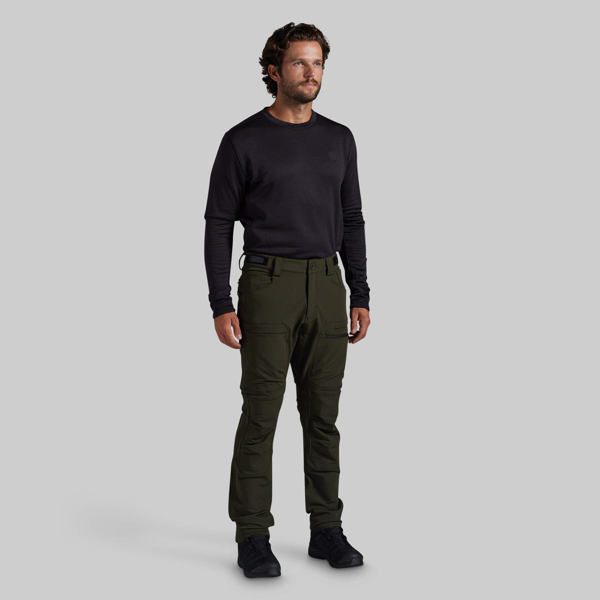 A man modeling olive green outdoor cargo pants with a black long-sleeve shirt and black boots, standing against a solid gray background.
