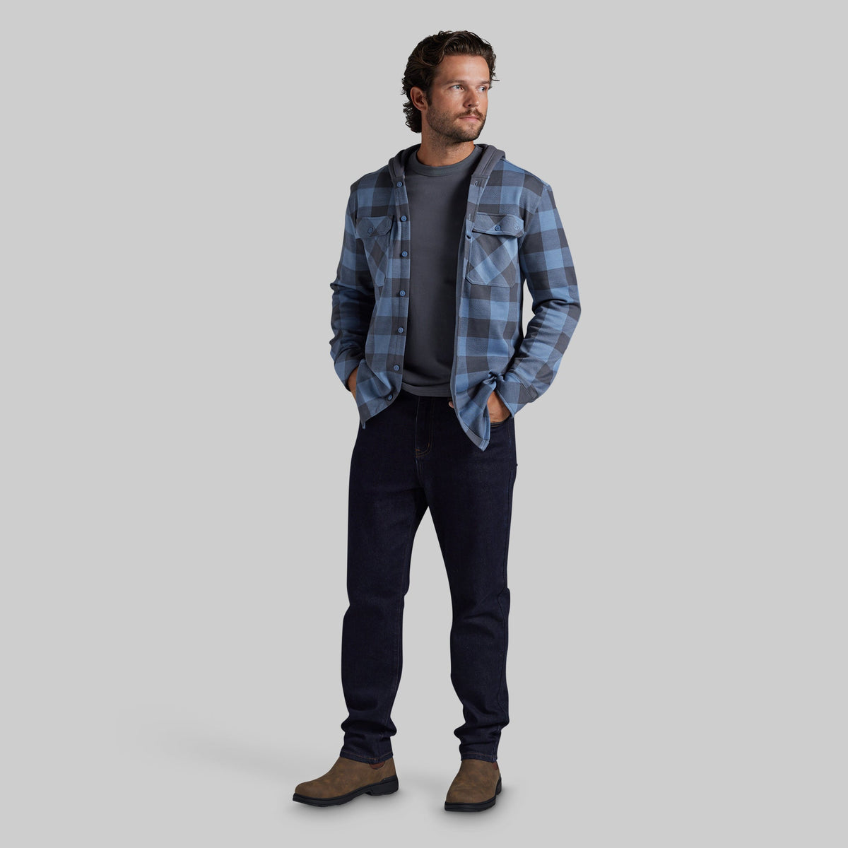 A man modeling a blue and gray plaid hooded flannel shirt over a dark gray t-shirt, dark jeans, and brown boots, standing against a solid gray studio background.