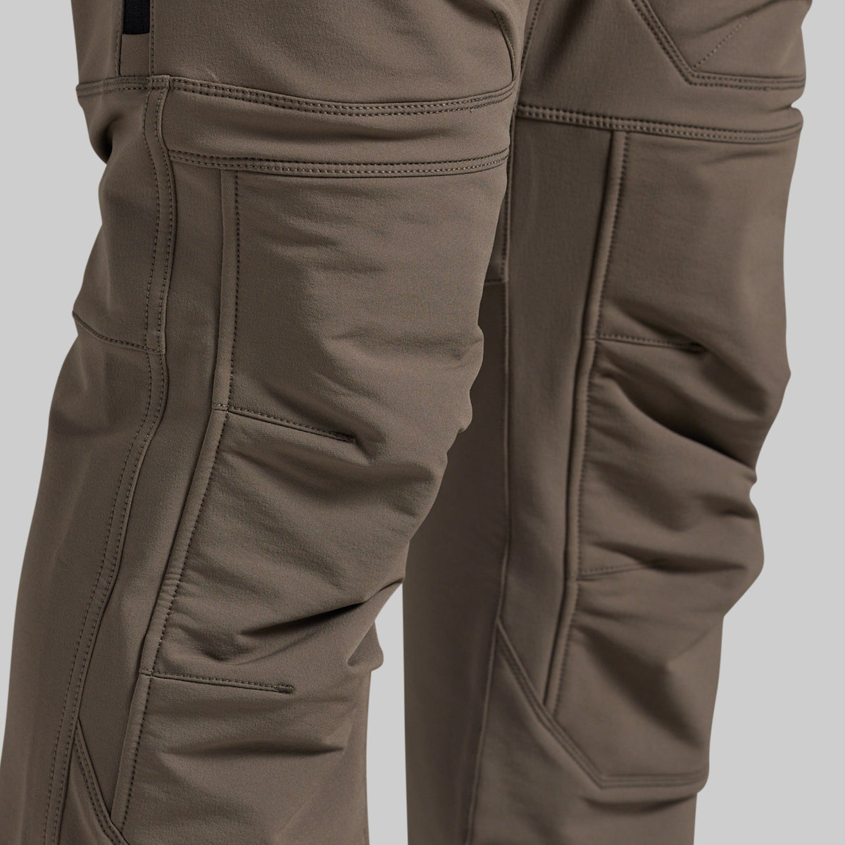 Close-up of a pair of technical outdoor pants in a warm grey color, showing the detailed stitching, reinforced knees, and fabric texture.