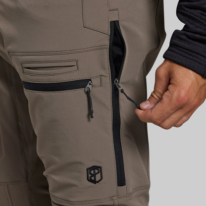 Close-up of a person wearing taupe-grey technical pants, showing a side zip pocket with a black zipper and a black logo patch near the lower leg, with a hand pulling the zipper tab.