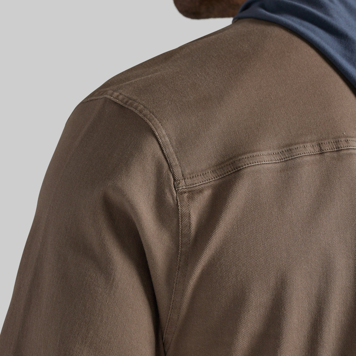 Close-up of a person wearing a brown textured overshirt or jacket, showing the shoulder seam and a blue shirt underneath, against a light gray background.
