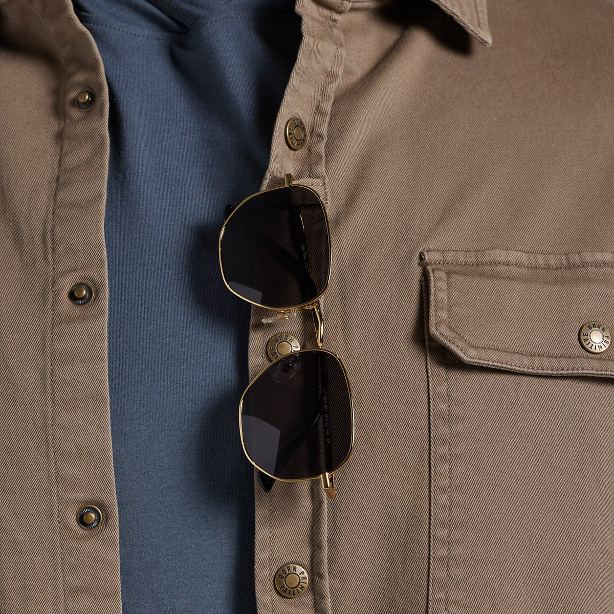 Close-up of sunglasses hanging from the placket of a tan overshirt worn over a blue shirt.