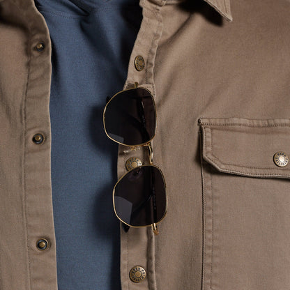 Close-up of sunglasses hanging from the placket of a tan overshirt worn over a blue shirt.