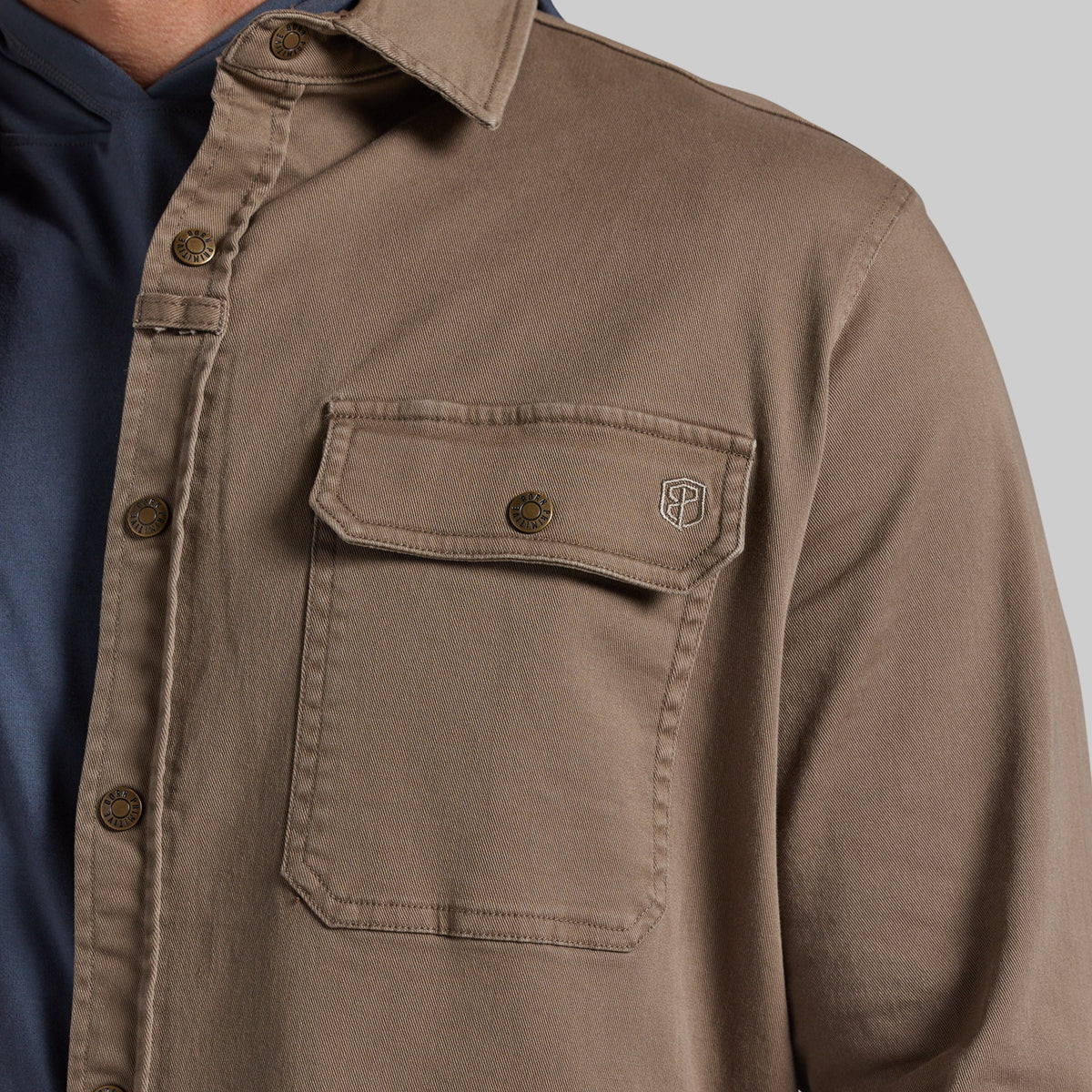 Close-up of a person wearing a brown button-front overshirt with a chest pocket and a small embroidered shield logo, layered over a navy shirt against a light gray background.
