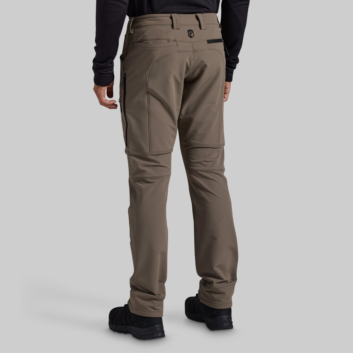 Rear view of a person wearing grey technical cargo pants with black boots and a black long-sleeve top, shown against a light gray studio background.