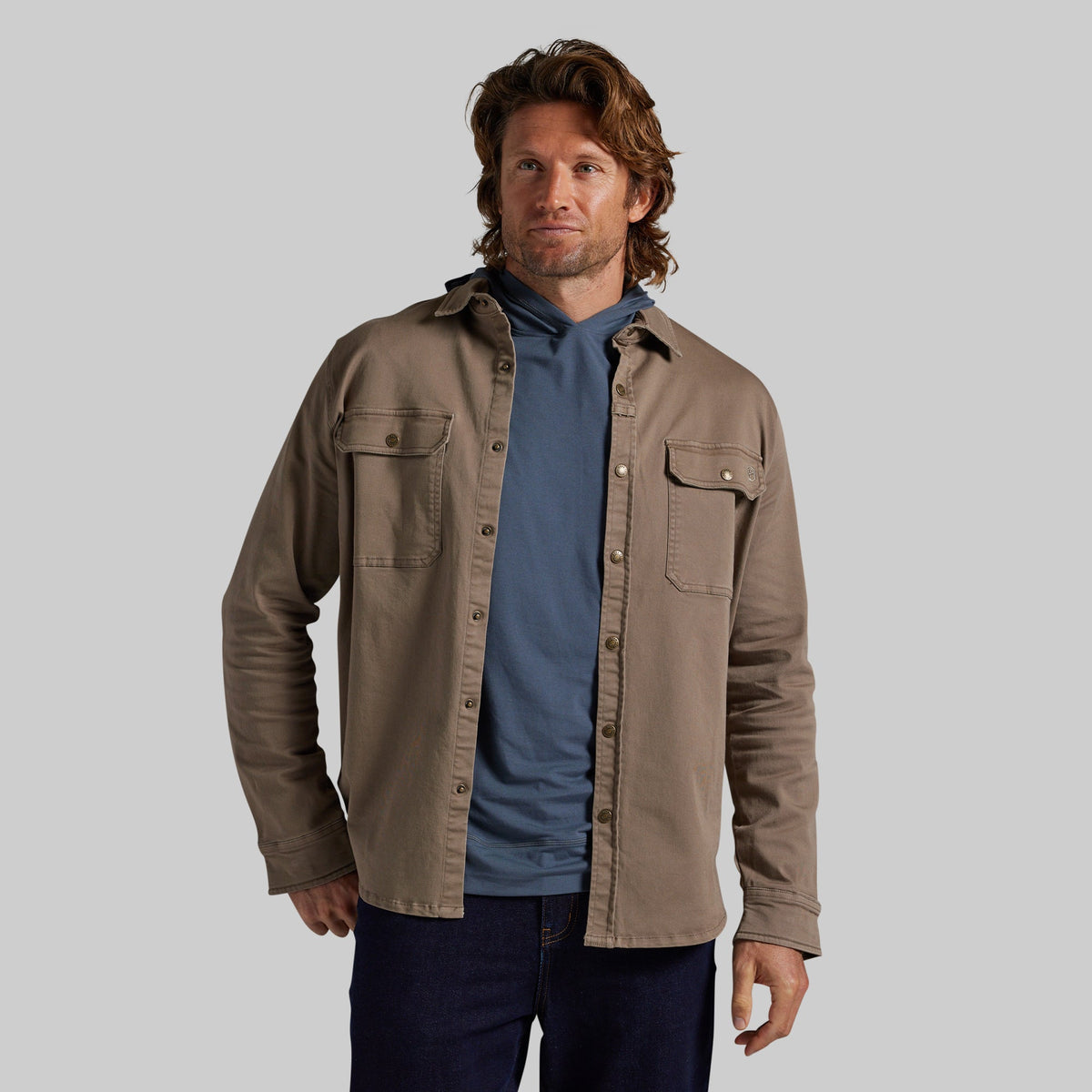 A man wearing a tan overshirt over a blue hoodie and dark jeans, standing against a solid gray studio background.