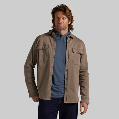 A man wearing a tan overshirt over a blue hoodie and dark jeans, standing against a solid gray studio background.