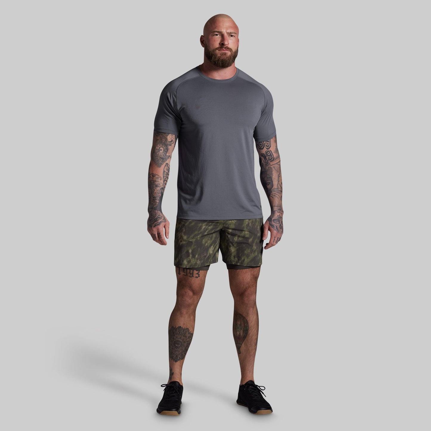 Man wearing a gray t-shirt and camouflage shorts on a plain background