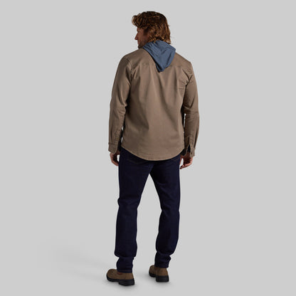 A man is shown from behind wearing a tan utility jacket, dark navy pants, and brown boots, photographed against a solid light gray background.