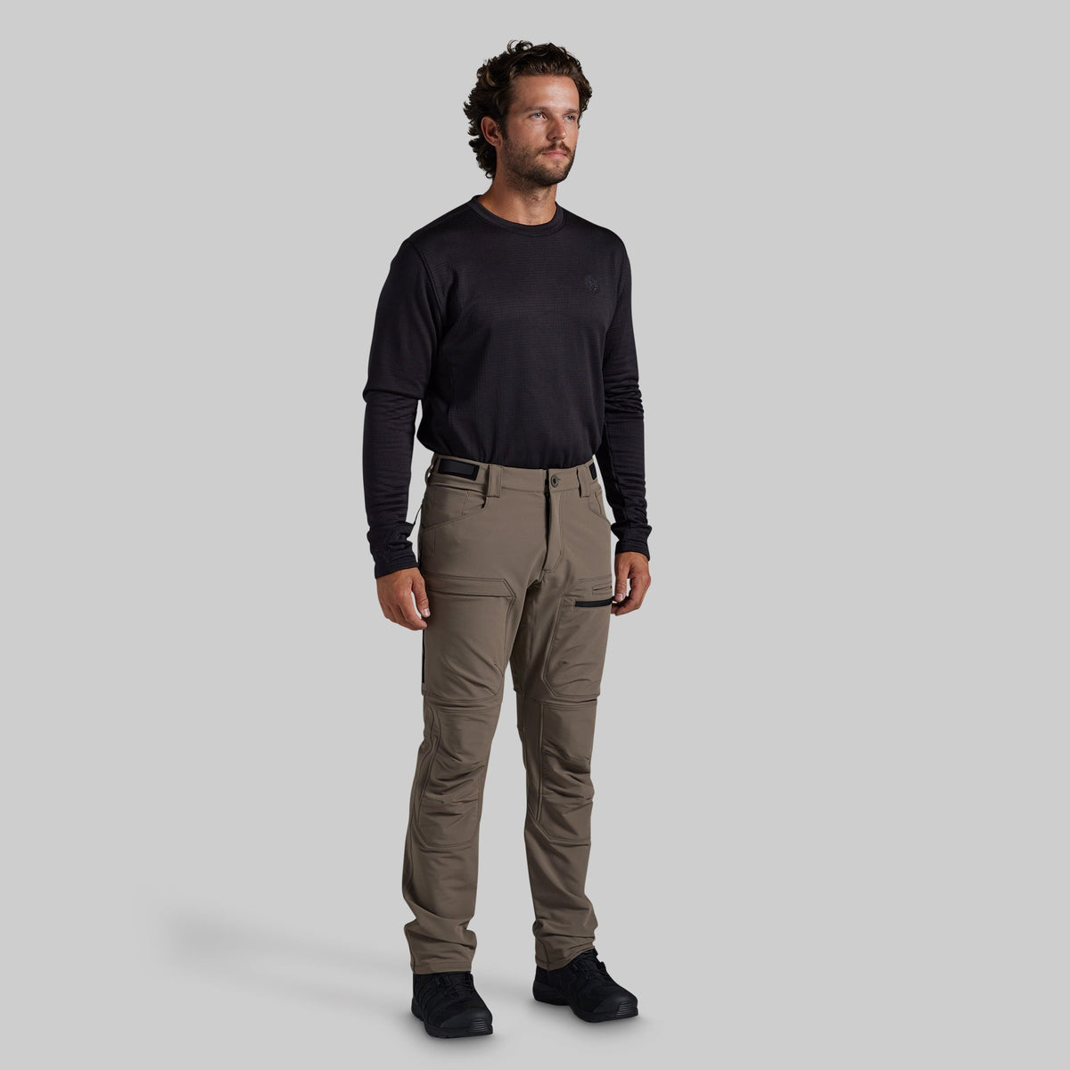 A man modeling a black long-sleeve shirt and grey technical cargo pants against a solid gray background.