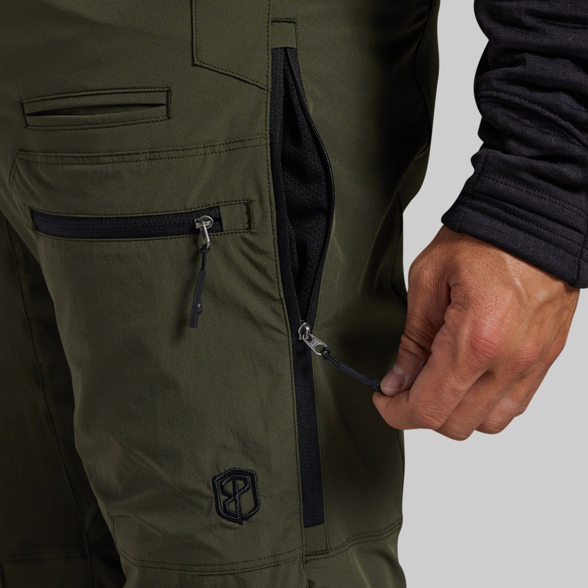 Close-up of olive green outdoor pants with a hand opening a black zipper pocket, showing the fabric texture and a small shield logo near the lower leg.