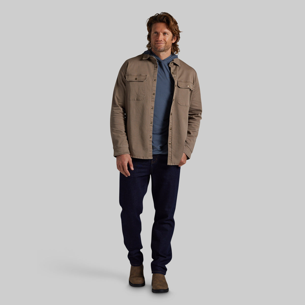 A man modeling a tan utility overshirt over a blue t-shirt, dark navy jeans, and brown boots, standing against a solid gray studio background.