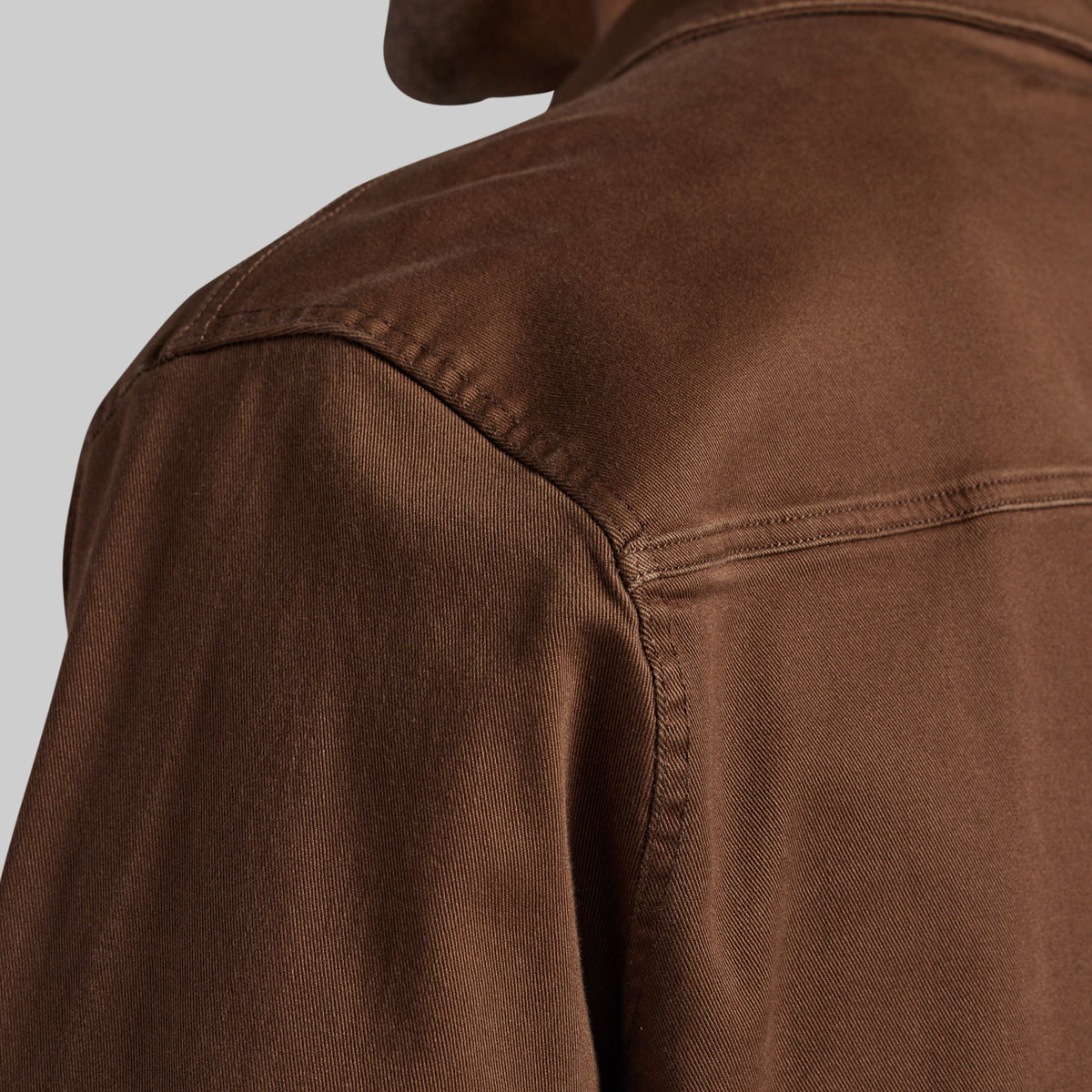 Close-up of a brown textured jacket or overshirt showing the shoulder seam, collar, and fabric texture against a light gray background.