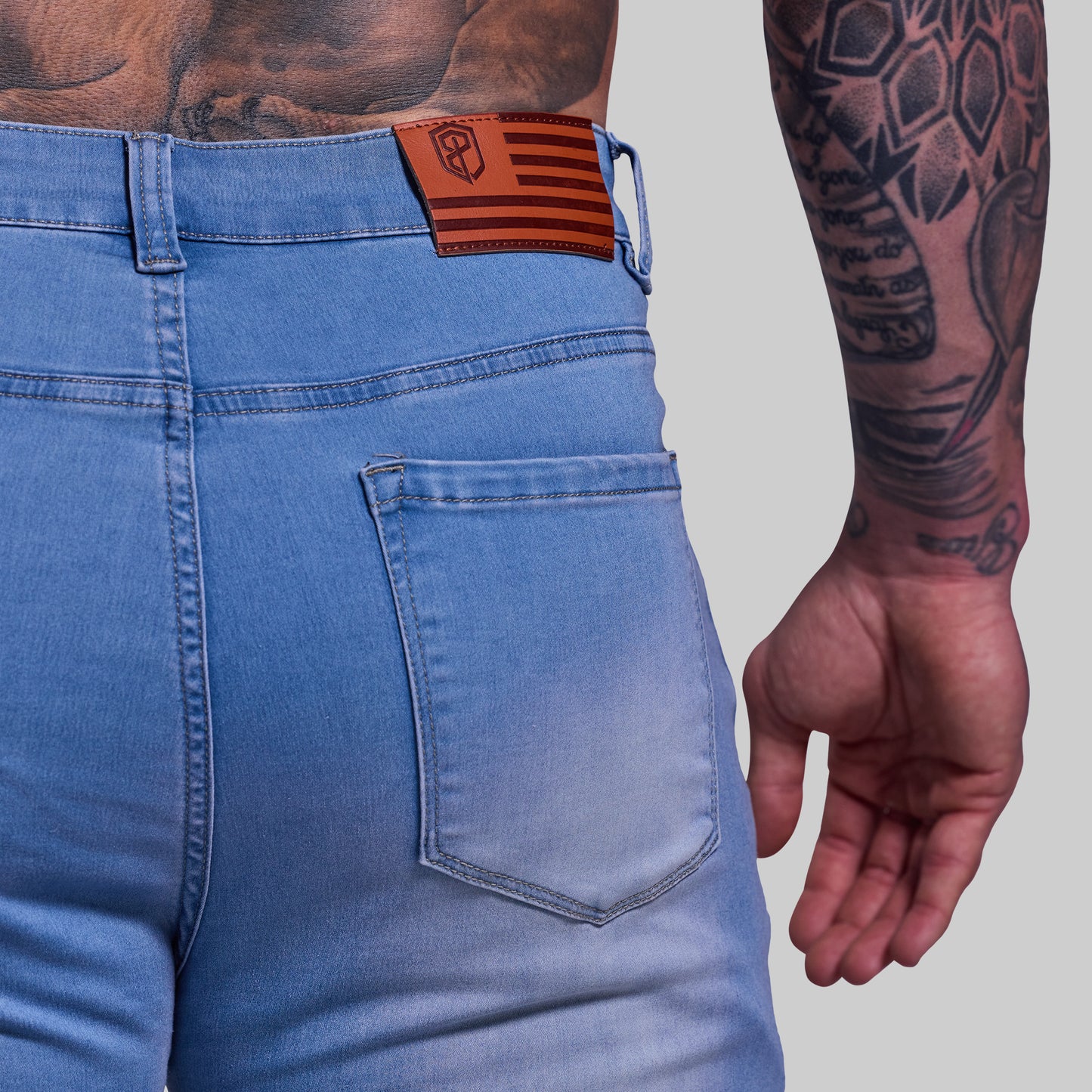 Blue jeans with a brown flag patch on the back pocket, worn by a person with tattoos.