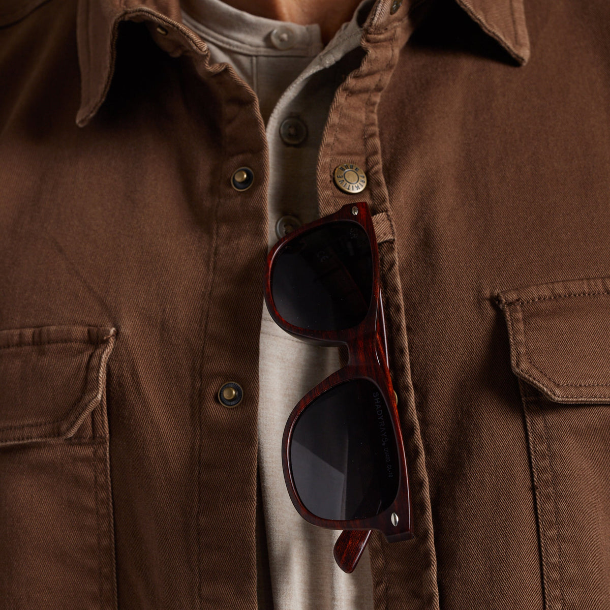 A close-up of a person wearing a brown overshirt over a light beige shirt, with dark sunglasses hanging from the sunglass loop.