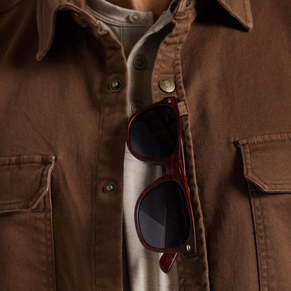 A close-up of a person wearing a brown overshirt over a light beige shirt, with dark sunglasses hanging from the sunglass loop.