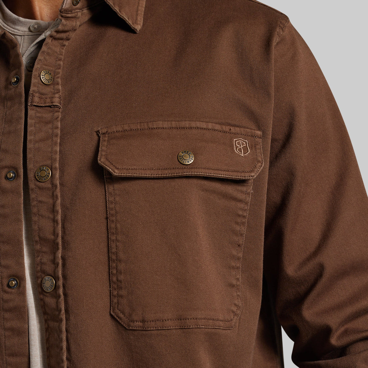 Close-up of a brown button-front shirt jacket with a chest pocket and a small embroidered logo, worn over a light gray shirt against a light gray background.
