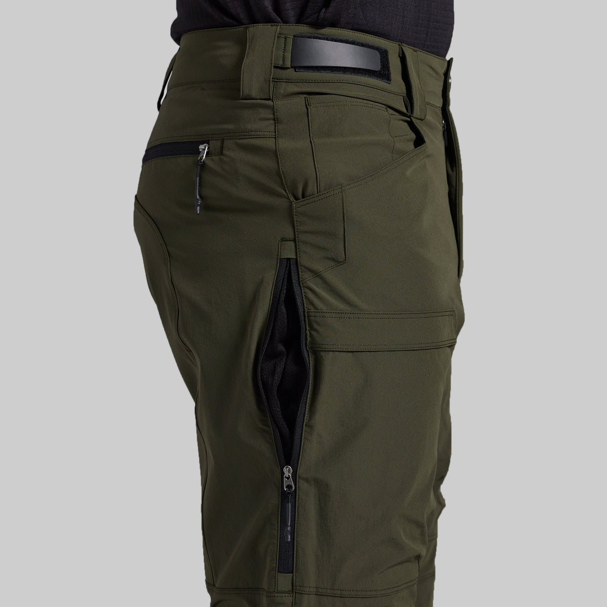 Close-up of olive green cargo pants with multiple zippered pockets, shown on a model from the waist down against a light gray background.