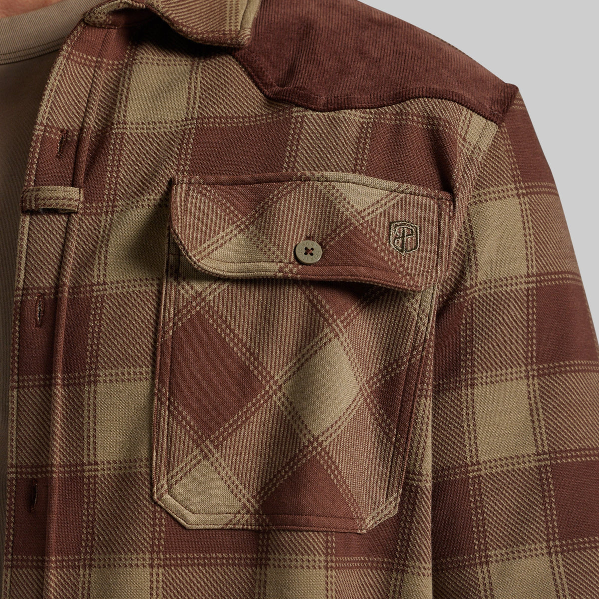 Close-up of a brown and beige plaid flannel shirt, showing a chest pocket with a button flap and a small shield logo patch.