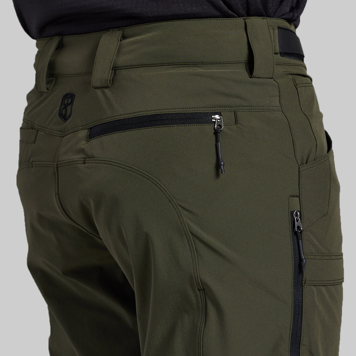 Close-up of the back waist and pockets of olive green technical outdoor pants, showing a zippered pocket, belt loops, and a small black logo patch.