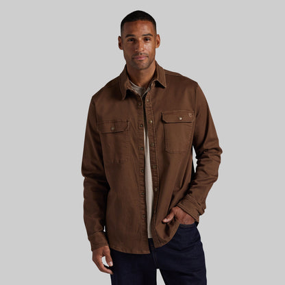 A man wearing a brown long-sleeve button-up shirt jacket over a light beige undershirt and dark blue jeans, standing against a solid gray background.