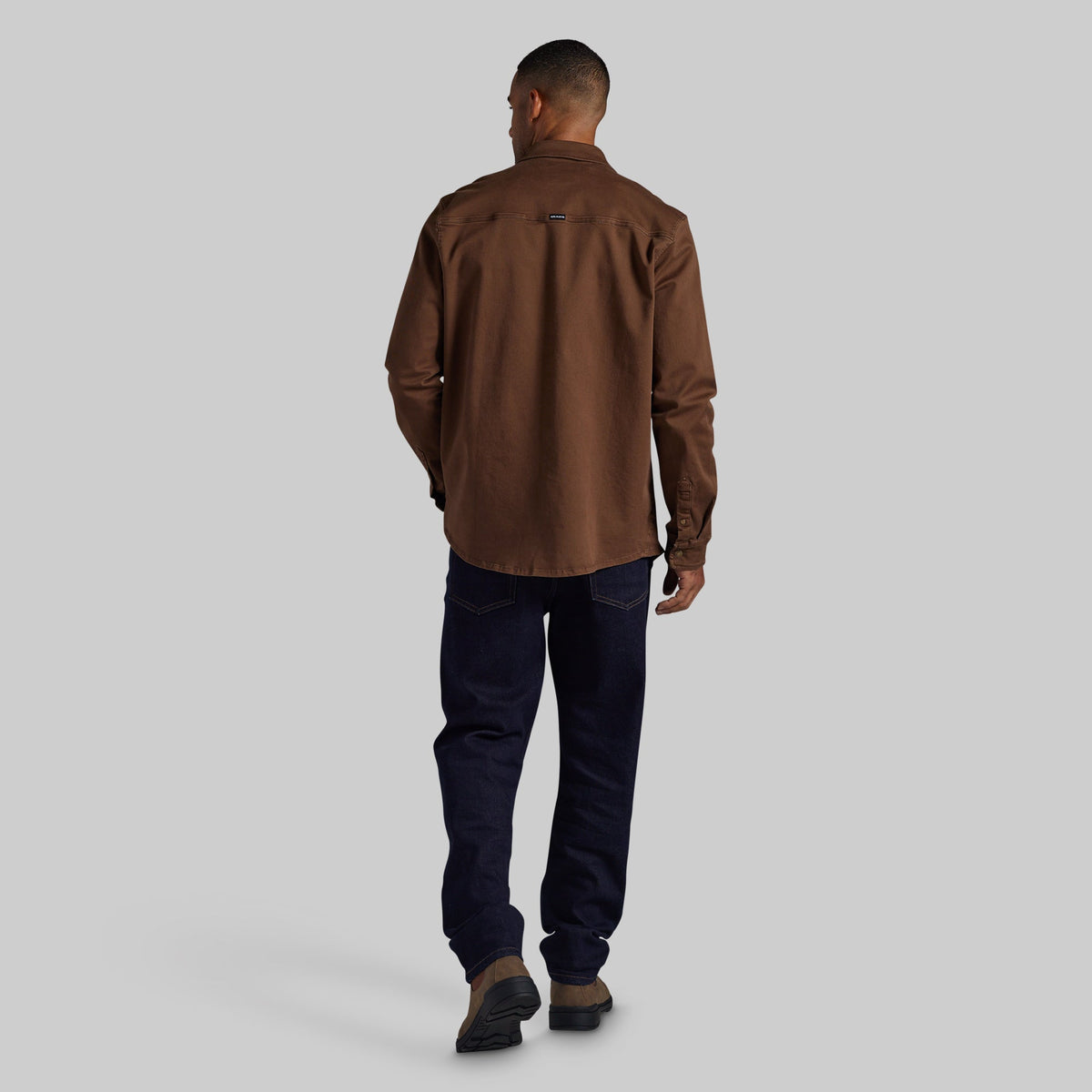 A man viewed from behind wearing a brown long-sleeve button-up shirt, dark navy jeans, and brown shoes, standing against a solid light gray background.