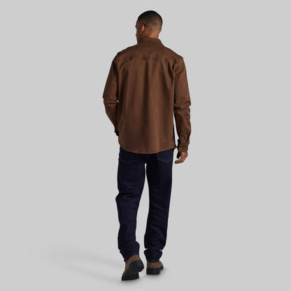 A man viewed from behind wearing a brown long-sleeve button-up shirt, dark navy jeans, and brown shoes, standing against a solid light gray background.