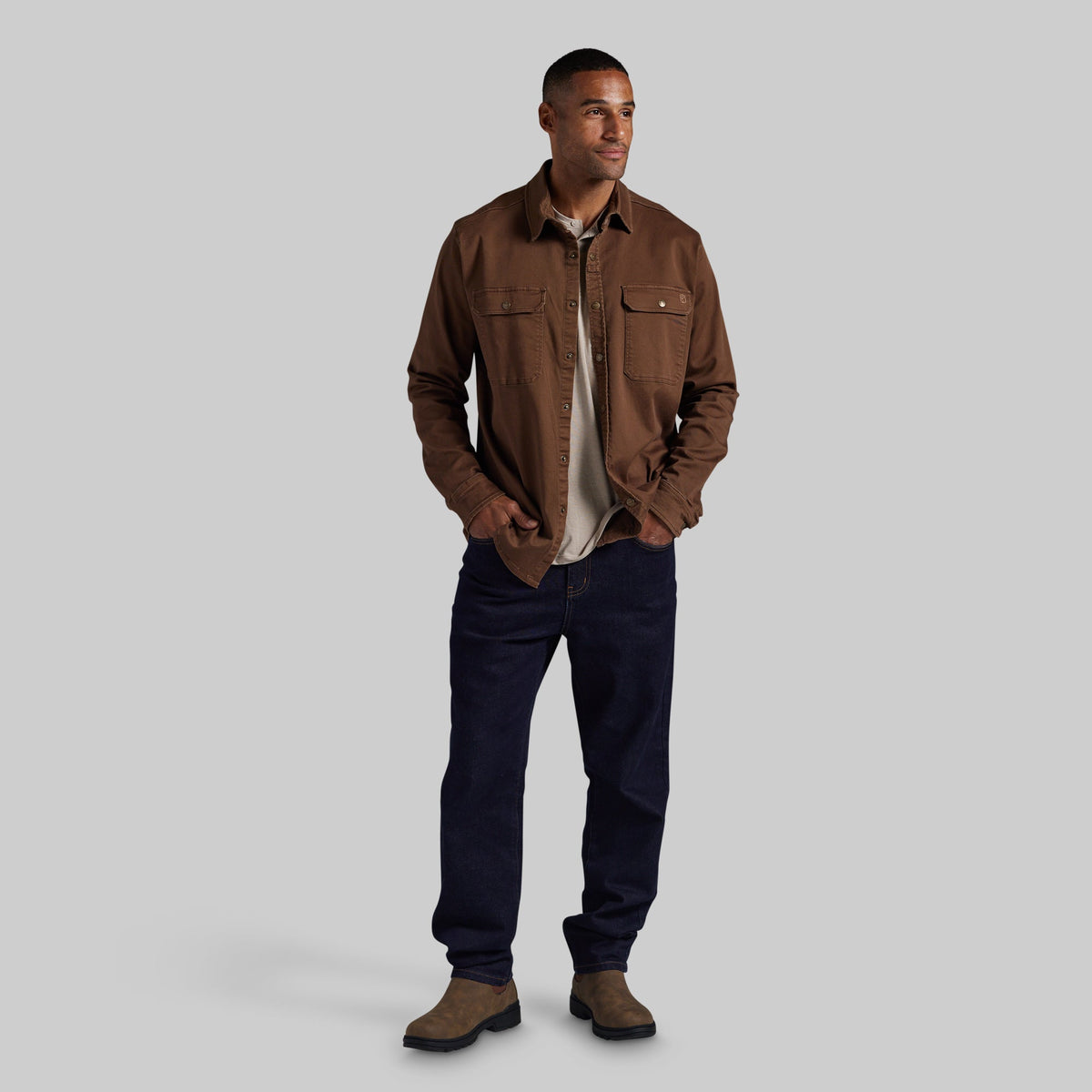 A man modeling a brown overshirt jacket over a light beige t-shirt, paired with dark navy jeans and brown suede boots, standing against a solid gray background.