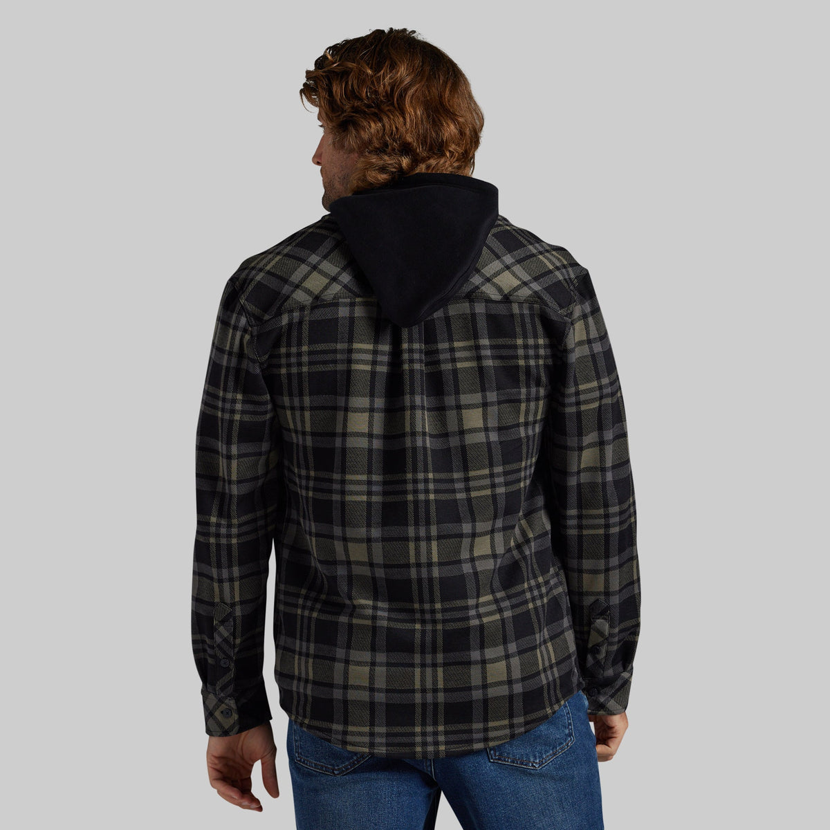 A man viewed from behind wearing a dark plaid hooded flannel with blue jeans, set against a solid gray studio background.