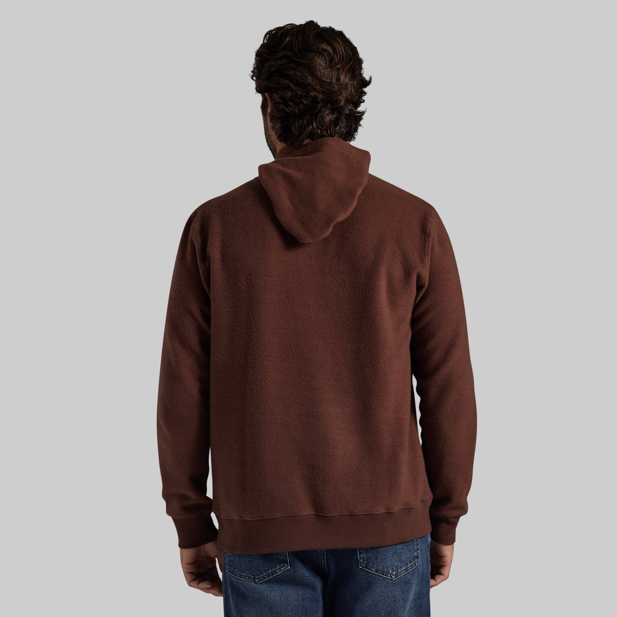 A man viewed from behind wearing a dark brown pullover hoodie and blue jeans, set against a solid light gray background.