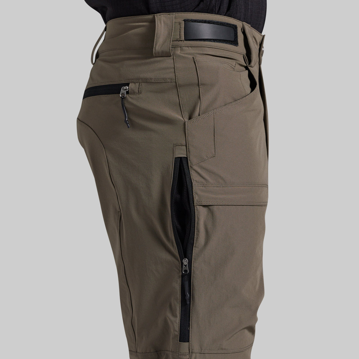 Close-up of a person wearing grey cargo pants with black zipper pockets and a black belt, shown against a light gray background.