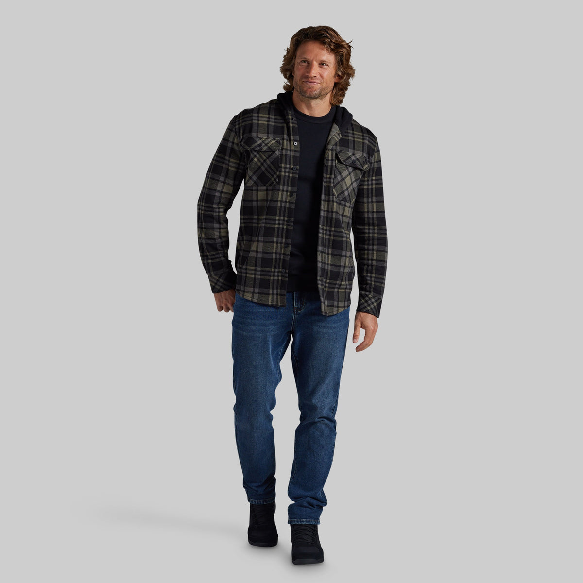 A man modeling a dark green and black plaid flannel shirt over a black undershirt, paired with blue jeans and black boots, against a solid gray background.